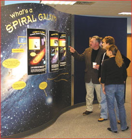 Three people stand in the right side of the image, looking at an exhibit at Space Place titled "What's a Spiral Galaxy?"